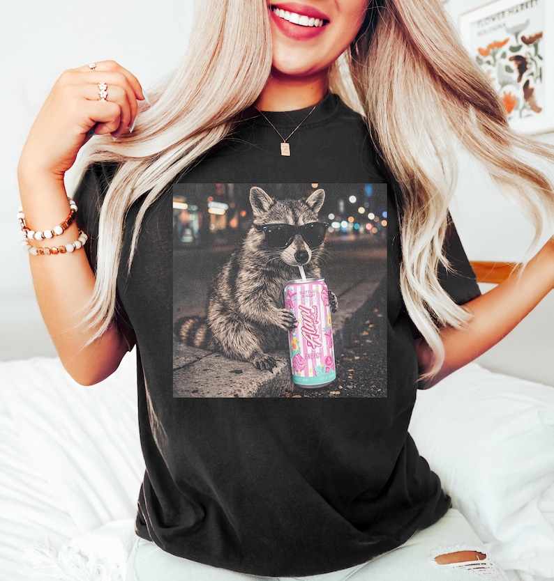 May include: Black t-shirt featuring a graphic of a raccoon wearing sunglasses and holding a pink can with the text "Fluff". The raccoon is sitting on a curb with a city background. The can has a floral and striped design.