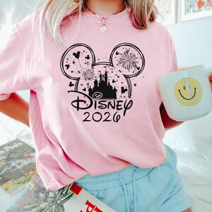 May include: A light pink t-shirt featuring a black graphic of Mickey Mouse ears with fireworks and a castle silhouette. The text "Disney 2026" is below the ears. The person is holding a white mug with a yellow smiley face.