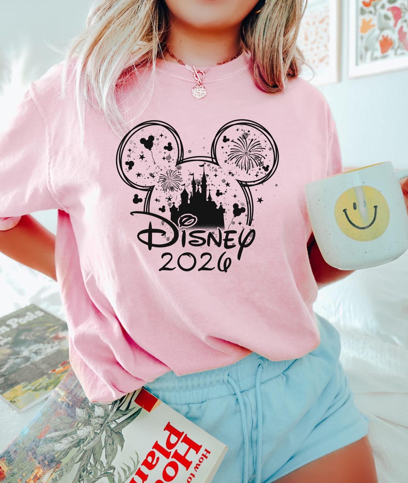 May include: A light pink t-shirt featuring a black graphic of Mickey Mouse ears with fireworks and a castle silhouette. The text "Disney 2026" is below the ears. The person is holding a white mug with a yellow smiley face.
