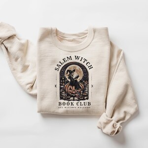 May include: Beige sweatshirt with a graphic featuring a witch on a pumpkin in front of a full moon. The text reads "Salem Witch Book Club" and "All Witches Welcome". The sweatshirt has long sleeves.