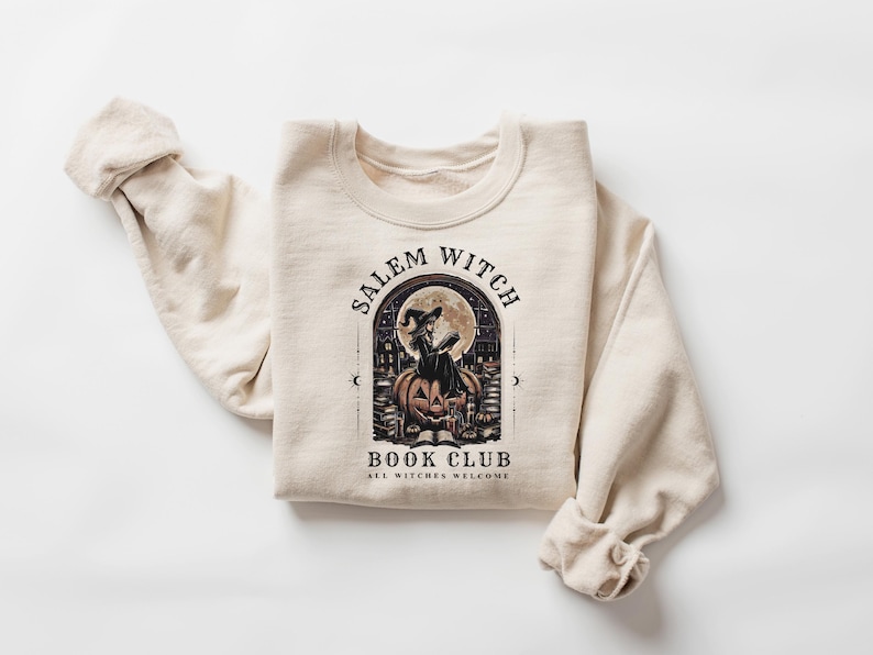 May include: Beige sweatshirt with a graphic featuring a witch on a pumpkin in front of a full moon. The text reads "Salem Witch Book Club" and "All Witches Welcome". The sweatshirt has long sleeves.