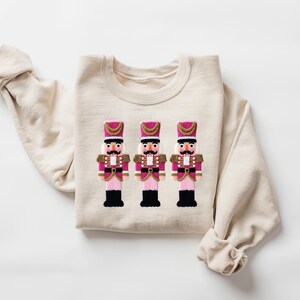 May include: A cream-colored sweatshirt featuring an embroidered design of three toy nutcracker soldiers. The nutcrackers have pink and gold accents, black boots, and white mustaches. The sweatshirt is folded with the sleeves visible.