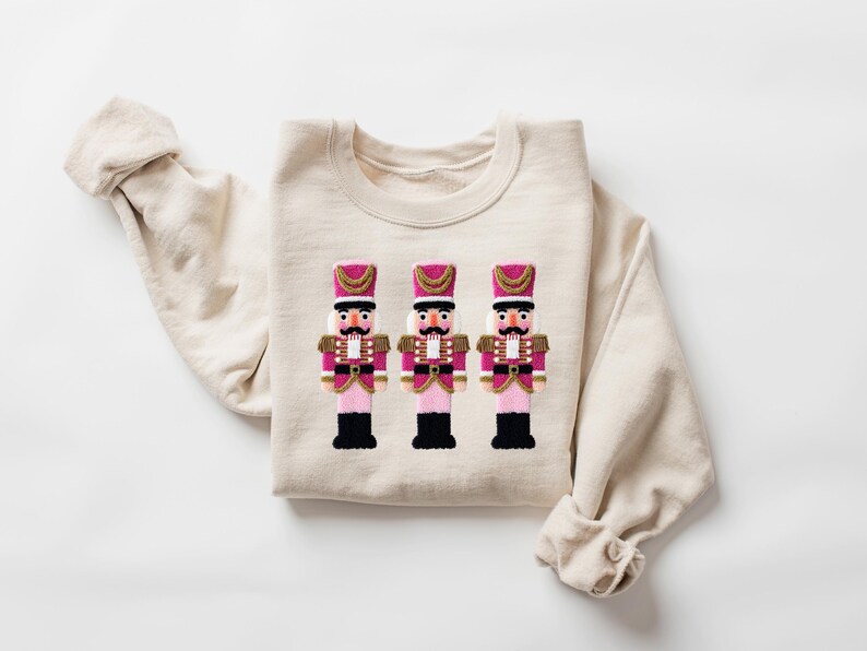 May include: A cream-colored sweatshirt featuring an embroidered design of three toy nutcracker soldiers. The nutcrackers have pink and gold accents, black boots, and white mustaches. The sweatshirt is folded with the sleeves visible.