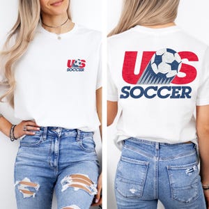 Puede incluir: Una camiseta blanca de manga corta con un peque&ntilde;o gr&aacute;fico en el pecho izquierdo con el texto US SOCCER y un bal&oacute;n de f&uacute;tbol. La parte posterior de la camiseta muestra un gr&aacute;fico grande con el texto US SOCCER en rojo y azul con un bal&oacute;n.