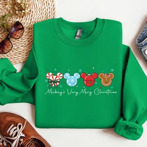 May include: A green crewneck sweatshirt featuring Mickey Mouse-themed Christmas ornaments. The ornaments include a candy cane, snowflake, red ornament, and gingerbread man design. The text "Mickey's Very Merry Christmas" is written in a script font.