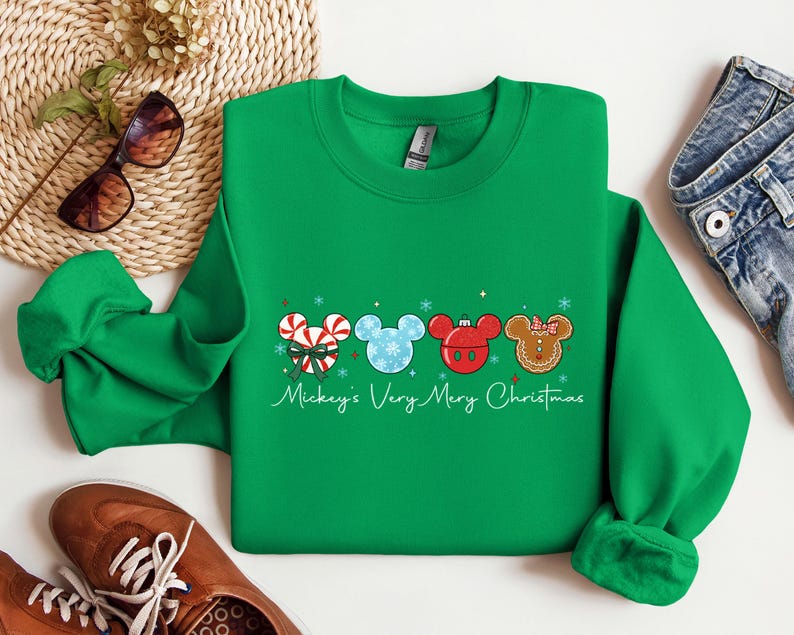 May include: A green crewneck sweatshirt featuring Mickey Mouse-themed Christmas ornaments. The ornaments include a candy cane, snowflake, red ornament, and gingerbread man design. The text "Mickey's Very Merry Christmas" is written in a script font.