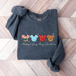 May include: A dark gray sweatshirt featuring a festive design with four Mickey Mouse-shaped ornaments. The ornaments include a candy cane, snowflake, red ornament, and gingerbread cookie. The text "Mickey's Very Merry Christmas" is written below the ornaments.
