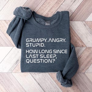 May include: A dark gray sweatshirt with the text "GRUMPY. ANGRY. STUPID. HOW LONG SINCE LAST SLEEP, QUESTION?" in white block letters. The sweatshirt is folded on a light brown and white wooden surface.