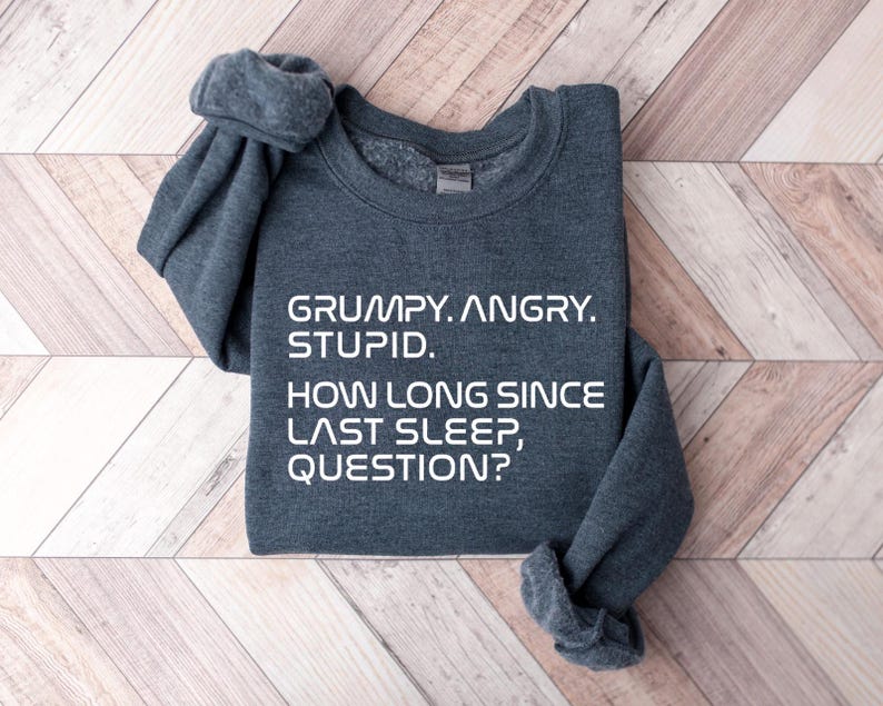 May include: A dark gray sweatshirt with the text "GRUMPY. ANGRY. STUPID. HOW LONG SINCE LAST SLEEP, QUESTION?" in white block letters. The sweatshirt is folded on a light brown and white wooden surface.