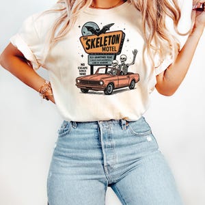 May include: Cream-colored t-shirt featuring a graphic of a motel sign that reads "The Skeleton Motel" with a bat and moon. Below the sign is a vintage car with two skeletons. The shirt also has the text "No Escape Since 1690".
