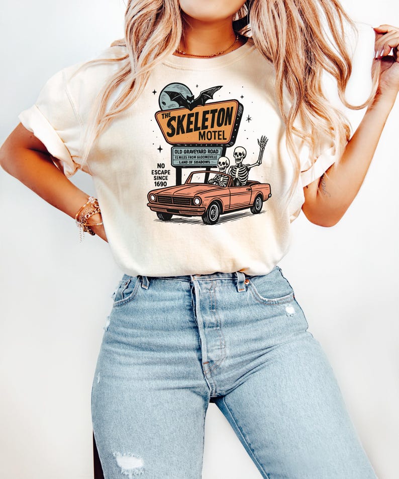 May include: Cream-colored t-shirt featuring a graphic of a motel sign that reads "The Skeleton Motel" with a bat and moon. Below the sign is a vintage car with two skeletons. The shirt also has the text "No Escape Since 1690".