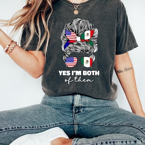 Może przedstawiać: Ciemnoszara koszulka z napisem "YES I'M BOTH of them". Grafika na koszulce przedstawia włosy kobiety stylizowane z flagami amerykańską i meksykańską. Kobieta nosi okulary przeciwsłoneczne z flagami amerykańską i meksykańską.