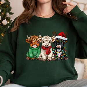 May include: Dark green sweatshirt with a Christmas design. It features three Highland cows, one in a green sweater, one with a red scarf, and one wearing a Santa hat and fairy lights. A festive winter theme.