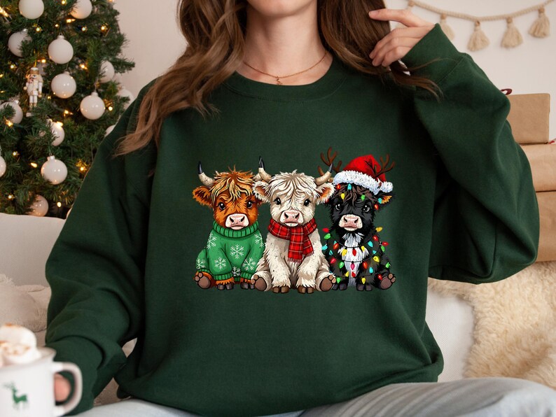 May include: Dark green sweatshirt with a Christmas design. It features three Highland cows, one in a green sweater, one with a red scarf, and one wearing a Santa hat and fairy lights. A festive winter theme.