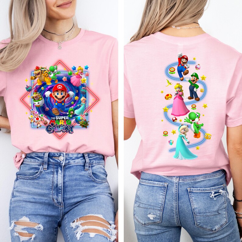 May include: A light pink t-shirt featuring a colorful graphic from the Super Mario Galaxy video game. The front design includes Mario, other characters, and the game's title. The back of the shirt shows Mario, Luigi, Princess Peach, Yoshi, and Elsa.