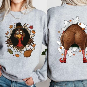 May include: A light gray sweatshirt featuring a Thanksgiving turkey design. The front shows a turkey wearing a pilgrim hat bursting through a torn hole, surrounded by fall leaves and pumpkins. The back shows the turkey's rear end, legs, and red shoes.