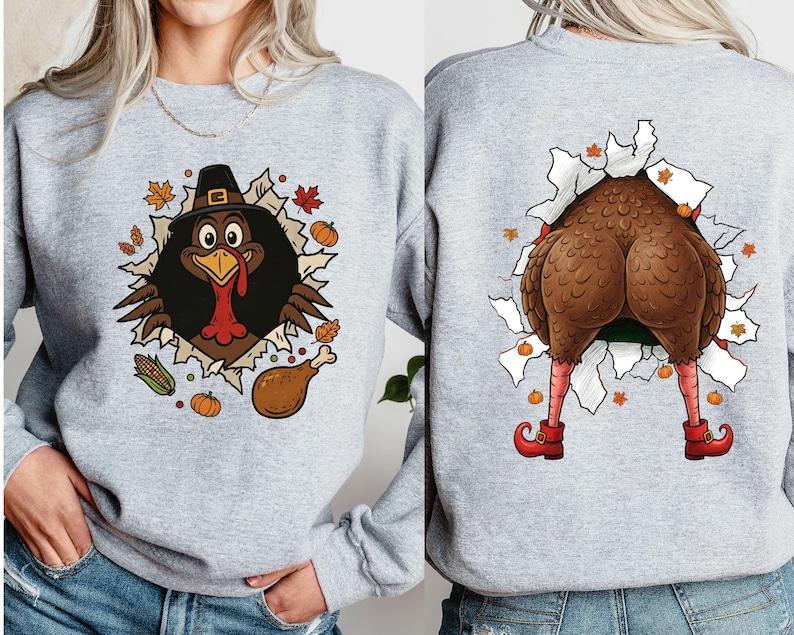 May include: A light gray sweatshirt featuring a Thanksgiving turkey design. The front shows a turkey wearing a pilgrim hat bursting through a torn hole, surrounded by fall leaves and pumpkins. The back shows the turkey's rear end, legs, and red shoes.
