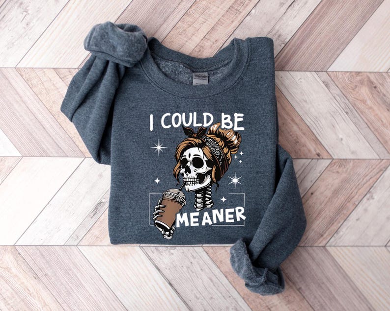 May include: Dark gray sweatshirt with the text "I COULD BE MEANER" and a graphic of a skeleton with a messy bun drinking from a coffee cup. The skeleton has a bandana and the design includes stars.