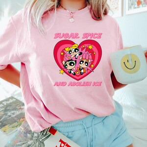 May include: A light pink t-shirt featuring a graphic of the Powerpuff Girls inside a heart with the text "SUGAR, SPICE AND ABOLISH ICE". The shirt is a crew neck style.