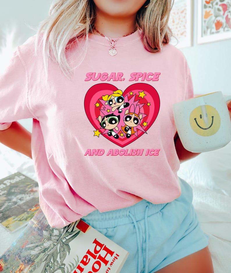 May include: A light pink t-shirt featuring a graphic of the Powerpuff Girls inside a heart with the text "SUGAR, SPICE AND ABOLISH ICE". The shirt is a crew neck style.