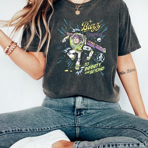 Op de afbeelding: Donkergrijs T-shirt met een afbeelding van Buzz Lightyear en de tekst "Buzz Lightyear" en "To Infinity and Beyond". Het shirt wordt gedragen met een blauwe jeans en witte sneakers. Het thema is een stripfiguur.
