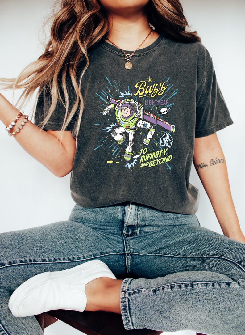 Op de afbeelding: Donkergrijs T-shirt met een afbeelding van Buzz Lightyear en de tekst "Buzz Lightyear" en "To Infinity and Beyond". Het shirt wordt gedragen met een blauwe jeans en witte sneakers. Het thema is een stripfiguur.