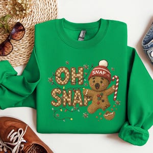 K&ouml;nnte beinhalten: Ein gr&uuml;nes Sweatshirt mit dem Schriftzug "OH SNAP" und einem Lebkuchenmann-Motiv. Der Lebkuchenmann tr&auml;gt eine rot-wei&szlig;e M&uuml;tze und h&auml;lt eine Zuckerstange. Das Sweatshirt hat einen Rundhalsausschnitt.