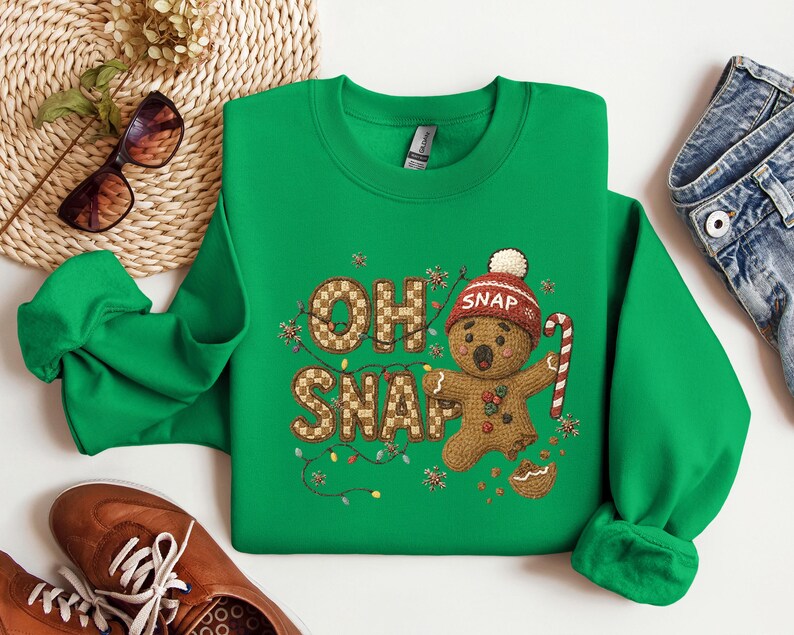K&ouml;nnte beinhalten: Ein gr&uuml;nes Sweatshirt mit dem Schriftzug "OH SNAP" und einem Lebkuchenmann-Motiv. Der Lebkuchenmann tr&auml;gt eine rot-wei&szlig;e M&uuml;tze und h&auml;lt eine Zuckerstange. Das Sweatshirt hat einen Rundhalsausschnitt.