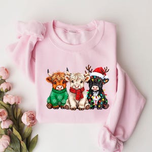 May include: A pale pink crew neck sweatshirt with a festive design. The graphic depicts three Highland cows, each wearing Christmas attire: a Santa hat, fairy lights, and festive sweaters. The design is centered.