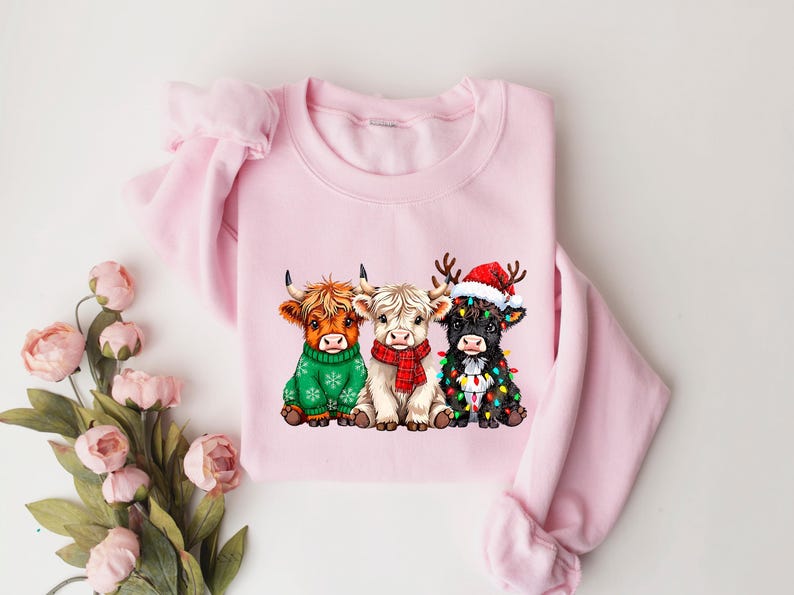 May include: A pale pink crew neck sweatshirt with a festive design. The graphic depicts three Highland cows, each wearing Christmas attire: a Santa hat, fairy lights, and festive sweaters. The design is centered.