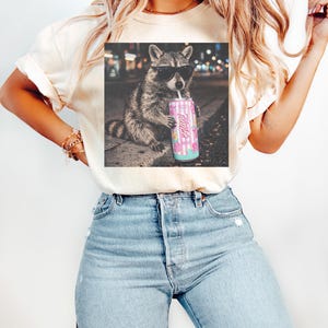 May include: Cream-colored t-shirt featuring a graphic of a raccoon wearing sunglasses and drinking from a pink can. The can has the word "Hi!" and a design of donuts and flowers. The raccoon is set against a blurred city background.