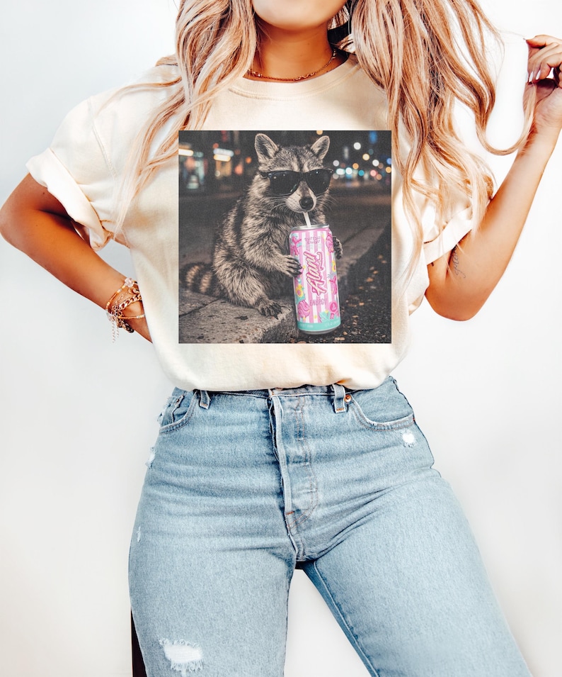 May include: Cream-colored t-shirt featuring a graphic of a raccoon wearing sunglasses and drinking from a pink can. The can has the word "Hi!" and a design of donuts and flowers. The raccoon is set against a blurred city background.