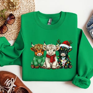 May include: A green sweatshirt with a festive design of three cartoon Highland cows. The cows are dressed for the holidays, wearing a green sweater, a red scarf, and a Santa hat with fairy lights. The sweatshirt is a vibrant green.
