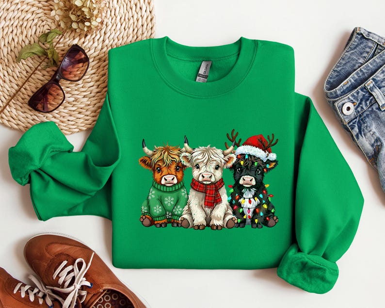 May include: A green sweatshirt with a festive design of three cartoon Highland cows. The cows are dressed for the holidays, wearing a green sweater, a red scarf, and a Santa hat with fairy lights. The sweatshirt is a vibrant green.