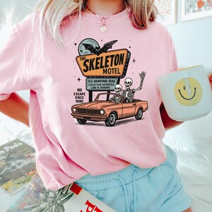 May include: Pink crewneck sweatshirt featuring a graphic of a vintage car with skeletons, a motel sign that reads "The Skeleton Motel", and the text "No Escape Since 1690". A white mug with a yellow smiley face is held in the hand.