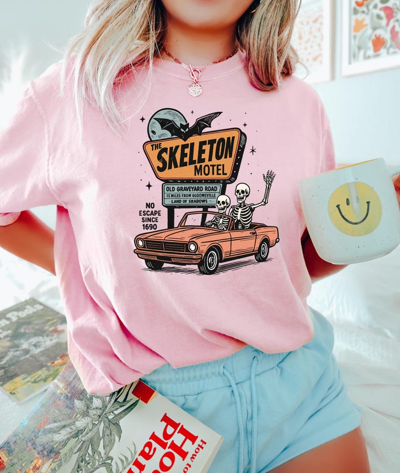 May include: Pink crewneck sweatshirt featuring a graphic of a vintage car with skeletons, a motel sign that reads "The Skeleton Motel", and the text "No Escape Since 1690". A white mug with a yellow smiley face is held in the hand.