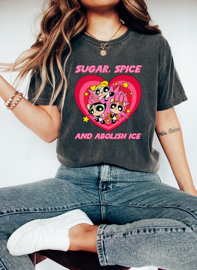 May include: Dark gray t-shirt featuring a pink heart with the Powerpuff Girls and the text "SUGAR. SPICE AND ABOLISH ICE". The shirt is a comfortable, casual style.