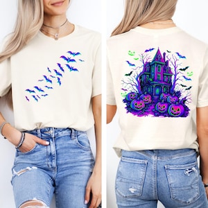 May include: Off-white t-shirt featuring a Halloween design. The front displays a flock of bats in shades of blue and purple. The back showcases a haunted house, pumpkins, and bats in vibrant colors, perfect for the spooky season.