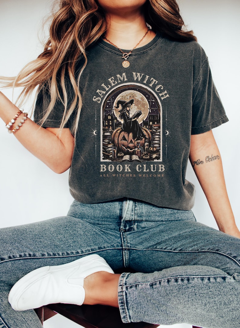May include: Dark gray t-shirt with a graphic featuring a witch reading on a pumpkin, with the text "Salem Witch Book Club, All Witches Welcome." The design includes a full moon and a gothic archway.