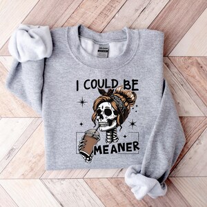 May include: A light gray sweatshirt featuring a skeleton with a bun and bandana, drinking from a coffee cup. The text "I COULD BE MEANER" is printed below the graphic. The sweatshirt has long sleeves and a crew neck.