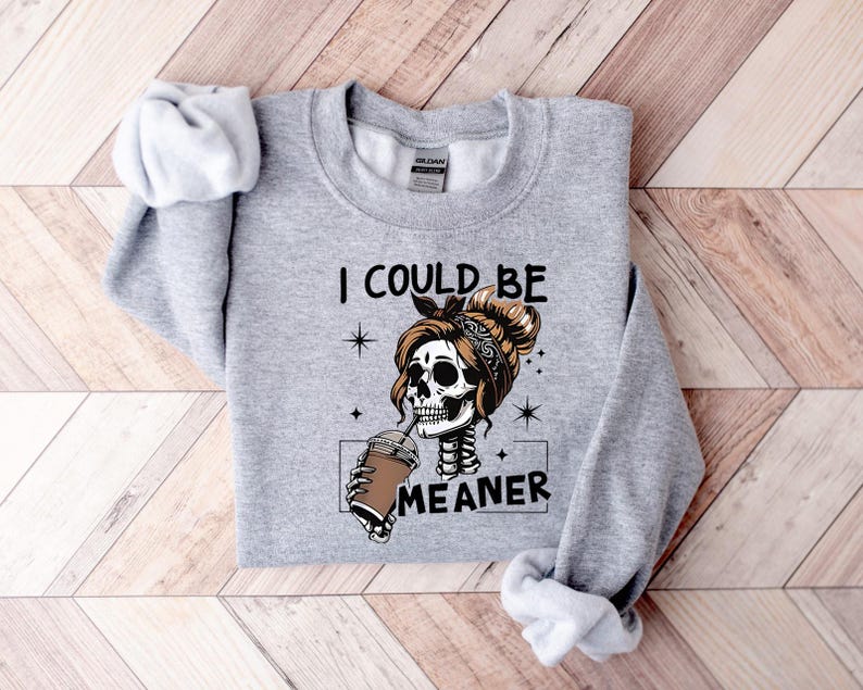 May include: A light gray sweatshirt featuring a skeleton with a bun and bandana, drinking from a coffee cup. The text "I COULD BE MEANER" is printed below the graphic. The sweatshirt has long sleeves and a crew neck.