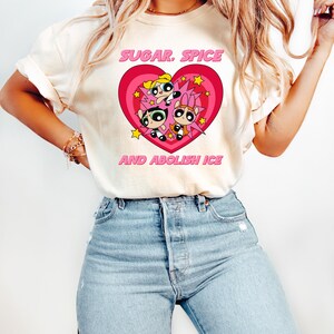 May include: Cream-colored t-shirt featuring a pink heart design with three cartoon characters and the text "SUGAR, SPICE AND ABOLISH ICE" in pink. The shirt is paired with blue jeans.