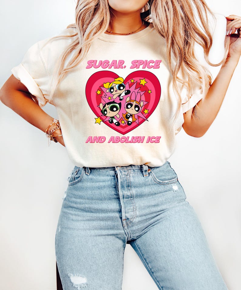 May include: Cream-colored t-shirt featuring a pink heart design with three cartoon characters and the text "SUGAR, SPICE AND ABOLISH ICE" in pink. The shirt is paired with blue jeans.