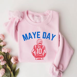 K&ouml;nnte beinhalten: Hellrosa Sweatshirt mit dem blauen Schriftzug "MAYE DAY" und einer roten Grafik eines Footballspielers mit der Nummer 10. Das Sweatshirt liegt gefaltet auf einer wei&szlig;en Oberfl&auml;che mit rosa Blumen.