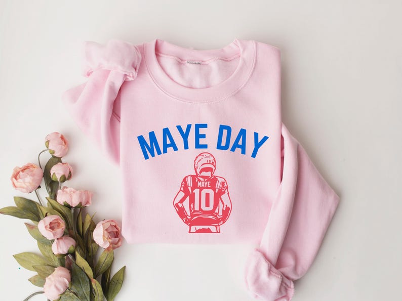 K&ouml;nnte beinhalten: Hellrosa Sweatshirt mit dem blauen Schriftzug "MAYE DAY" und einer roten Grafik eines Footballspielers mit der Nummer 10. Das Sweatshirt liegt gefaltet auf einer wei&szlig;en Oberfl&auml;che mit rosa Blumen.