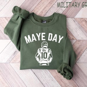 K&ouml;nnte beinhalten: Milit&auml;rgr&uuml;nes Sweatshirt mit dem wei&szlig;en Schriftzug "MAYE DAY". Darunter befindet sich eine wei&szlig;e Grafik eines Footballspielers mit dem Namen "MAYE" und der Nummer "10". Das Sweatshirt hat einen Rundhalsausschnitt und lange &Auml;rmel.