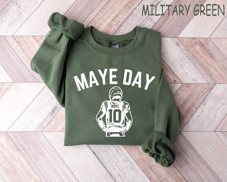 K&ouml;nnte beinhalten: Milit&auml;rgr&uuml;nes Sweatshirt mit dem wei&szlig;en Schriftzug "MAYE DAY". Darunter befindet sich eine wei&szlig;e Grafik eines Footballspielers mit dem Namen "MAYE" und der Nummer "10". Das Sweatshirt hat einen Rundhalsausschnitt und lange &Auml;rmel.