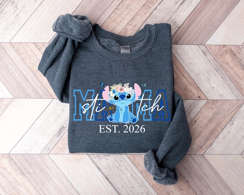 May include: A dark gray sweatshirt featuring a blue cartoon character with a floral crown and the text "Mama Stitch EST. 2026". The design includes blue butterflies and lettering.