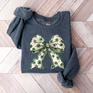 May include: A dark teal sweatshirt featuring a large bow design. The bow has a cream-colored background with green shamrocks and green trim. The sweatshirt is folded on a light wood-grain surface.