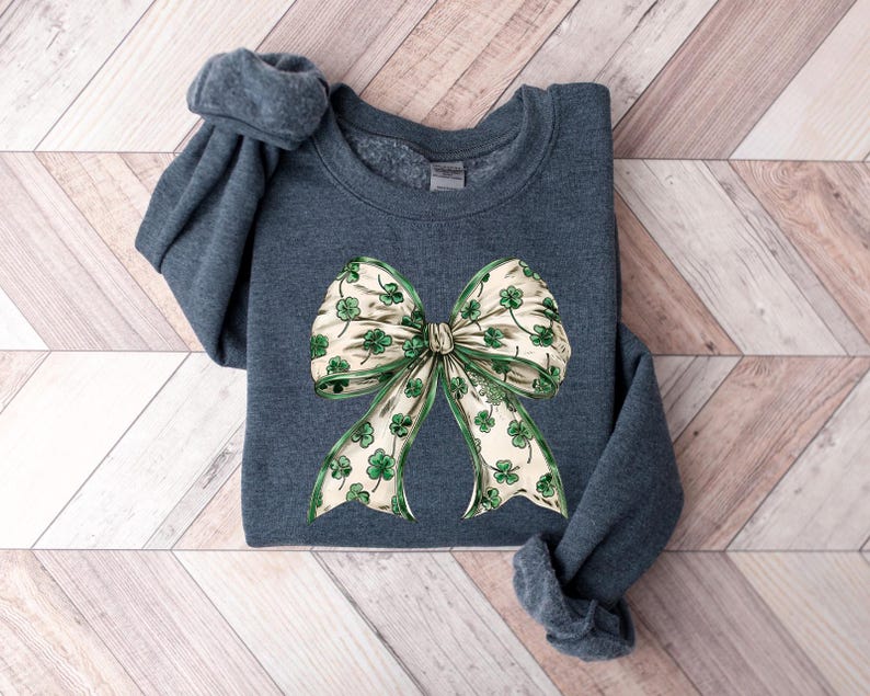 May include: A dark teal sweatshirt featuring a large bow design. The bow has a cream-colored background with green shamrocks and green trim. The sweatshirt is folded on a light wood-grain surface.